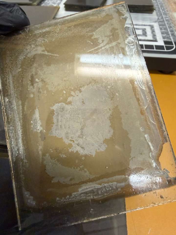 A small photographic glass plate being held on the edges by a gloved hand. The emulsion on the plate shows severe signs of degradation, flaking, and discoloration. An image is not immediately apparent.
