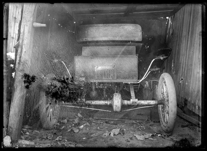 A scan of a glass negative showing the back of a very old car inside some sort of barn, shed, or garage. Neither the car nor the shed looks to be in very good shape. The license plate says 'PENNA 1908' above the license plate number: 14183. The large area of paper has been removed, revealing a bit more of the image and a smaller area where the image has been completely lost.