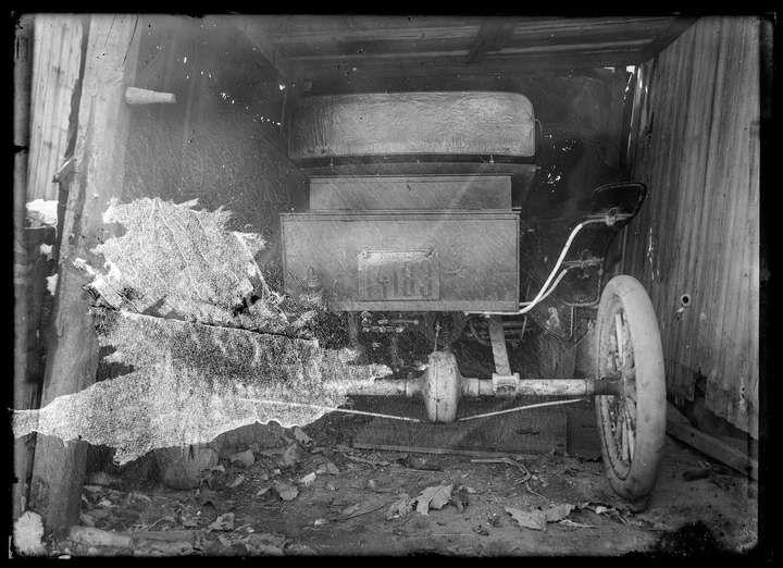 A scan of a glass negative showing the back of a very old car inside some sort of barn, shed, or garage. Neither the car nor the shed looks to be in very good shape. The license plate says 'PENNA 1908' above the license plate number: 14183. There is large area of the image that cannot be seen because of a fibrous paper that is stuck to the emulsion.